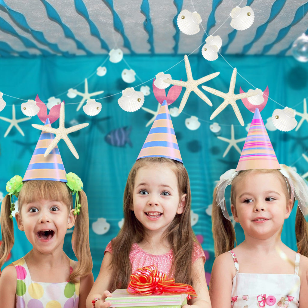 Three young girls wearing party hats and smiling, with seashell and starfish garlands in the background.