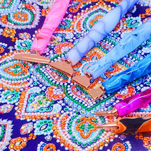 Four diamond painting pens with rose gold tips resting on a colorful patterned canvas with rhinestones.