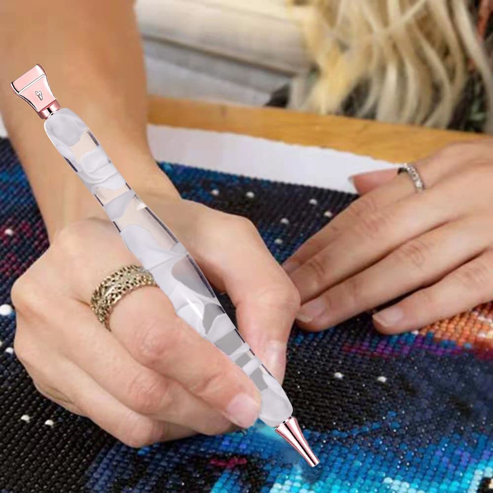 Close-up of a person's hands using a white rose gold diamond painting pen on a sparkling canvas. The pen has a marbled white and clear barrel.