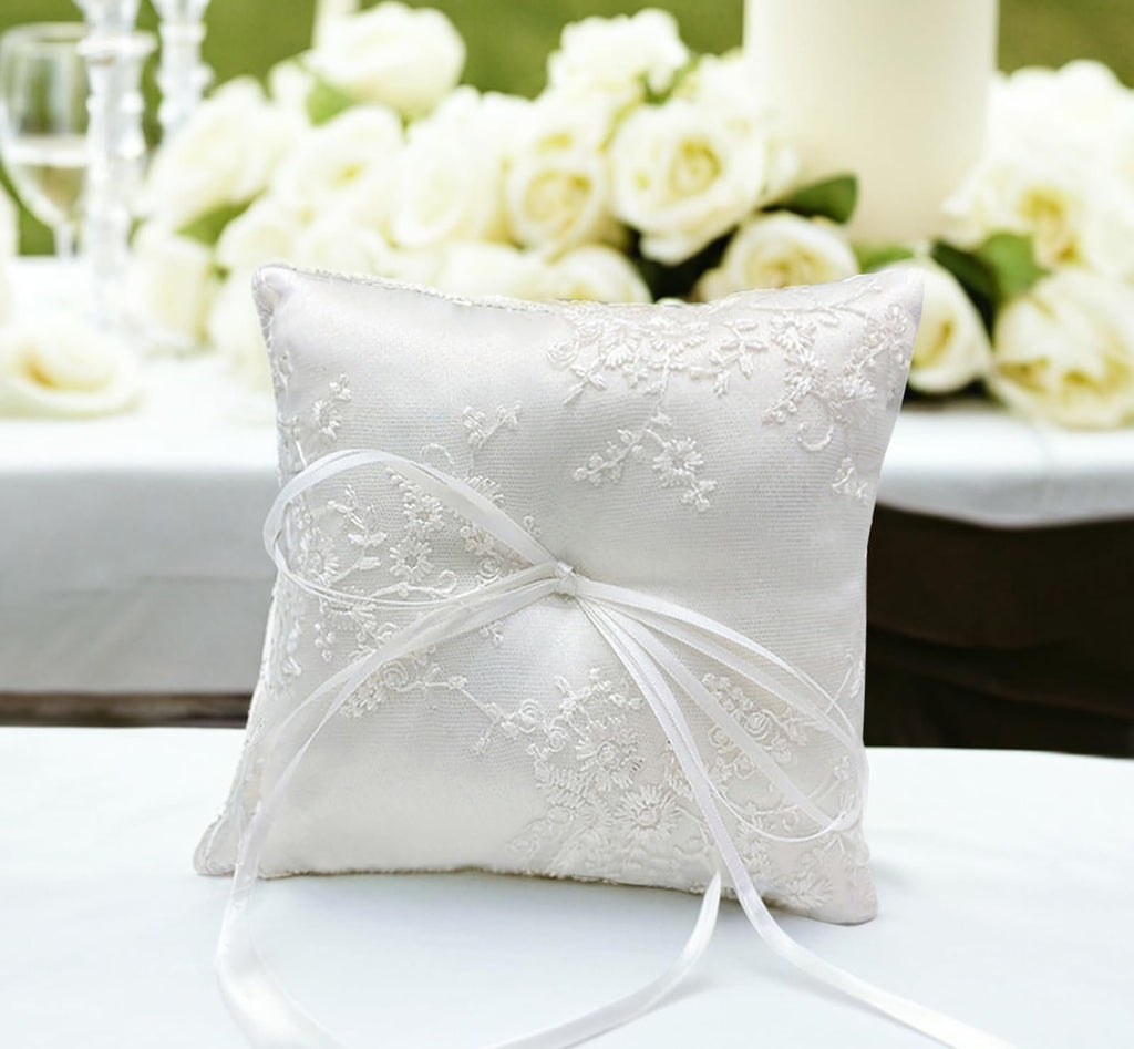 White satin ring bearer pillow with floral lace embroidery and ribbon bow, set on a white surface with blurred wedding decor in the background.
