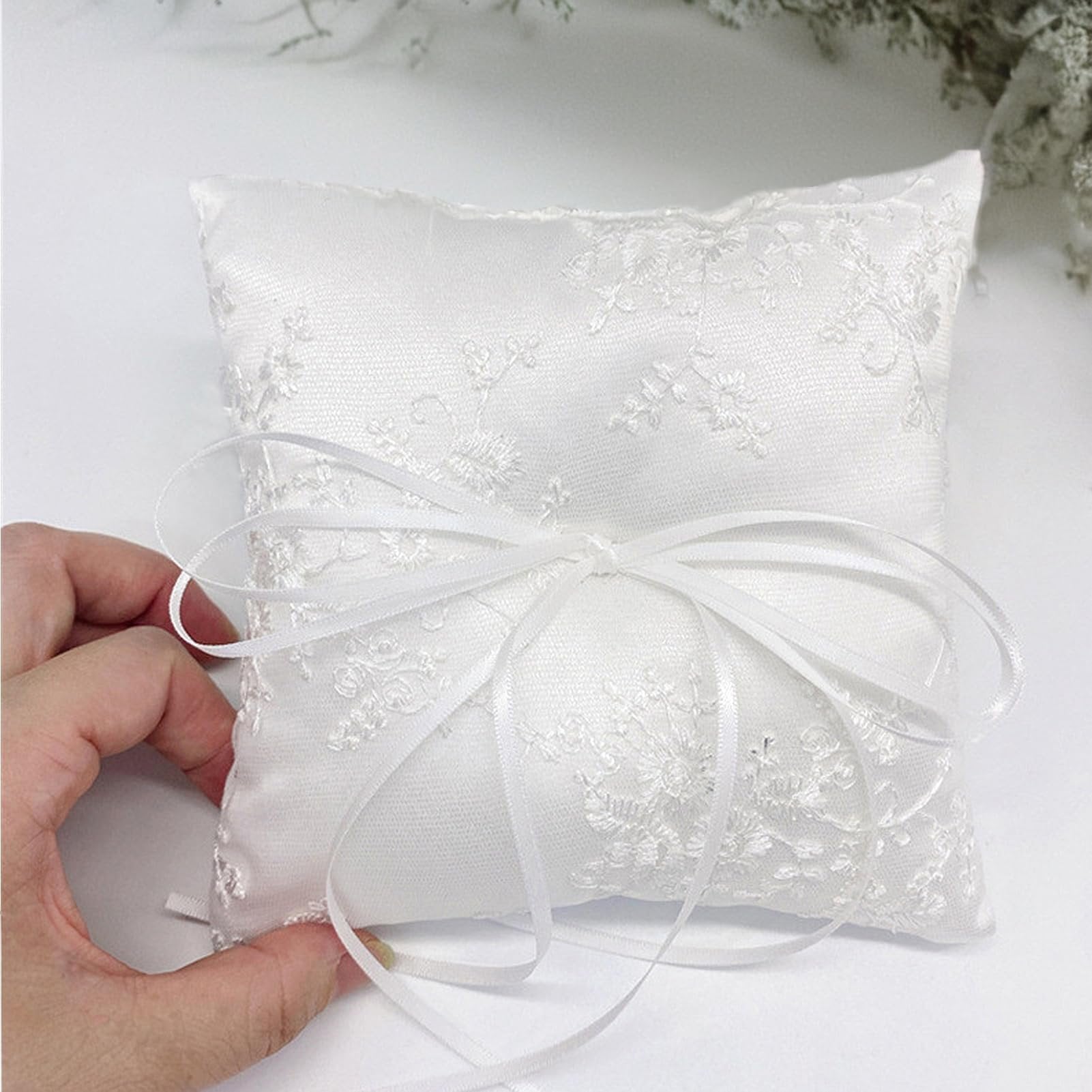 White satin ring bearer pillow with floral lace embroidery and ribbon bow, held by a hand.