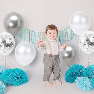 Baby boy in suspenders surrounded by white, silver, and confetti balloons and teal pom-poms