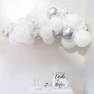White and silver confetti balloons arranged in a garland on a white wall, with a 'Cards & Wishes' box on a table below.