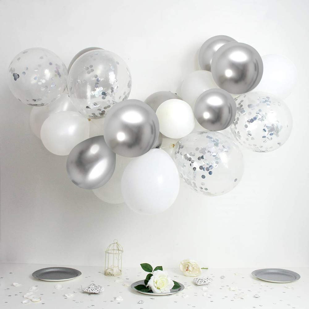 Assortment of white, silver, and confetti balloons arranged in a cluster against a white background, with decorative elements on a table below.