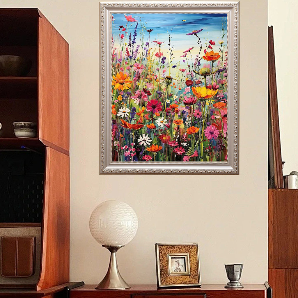 Framed painting of a vibrant field of wildflowers with birds in a blue sky, displayed on a wall above a table with a lamp and picture frame.