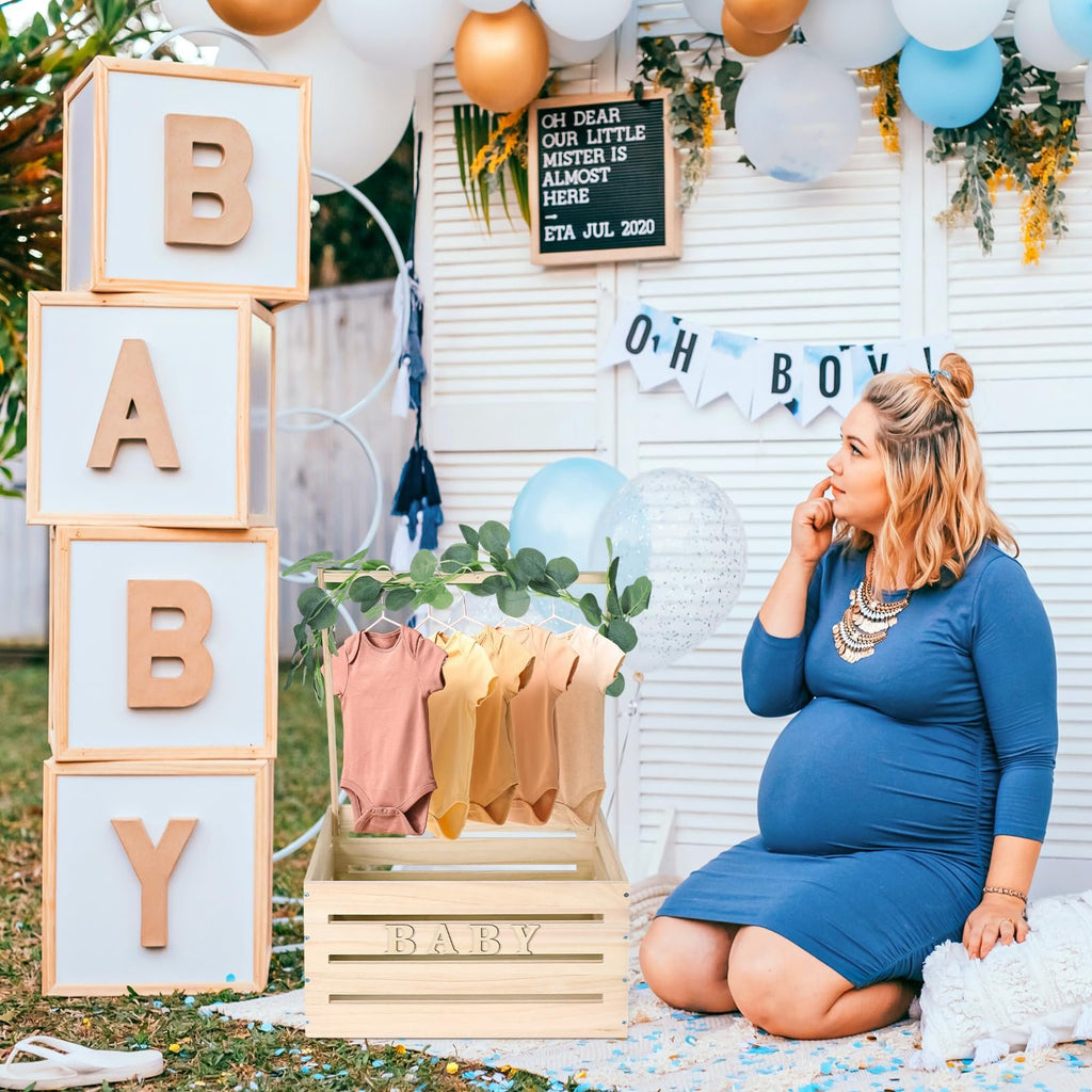 Pregnant woman at a baby shower with a wooden crate displaying baby onesies and large letter blocks spelling BABY.