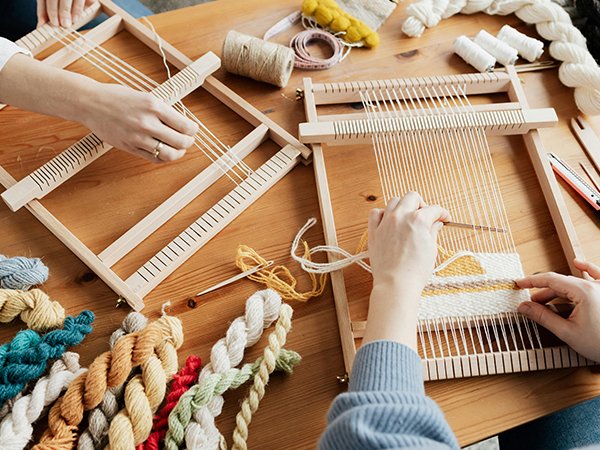 Two people weaving on wooden tapestry frames with colorful yarn and tools on a wooden table.