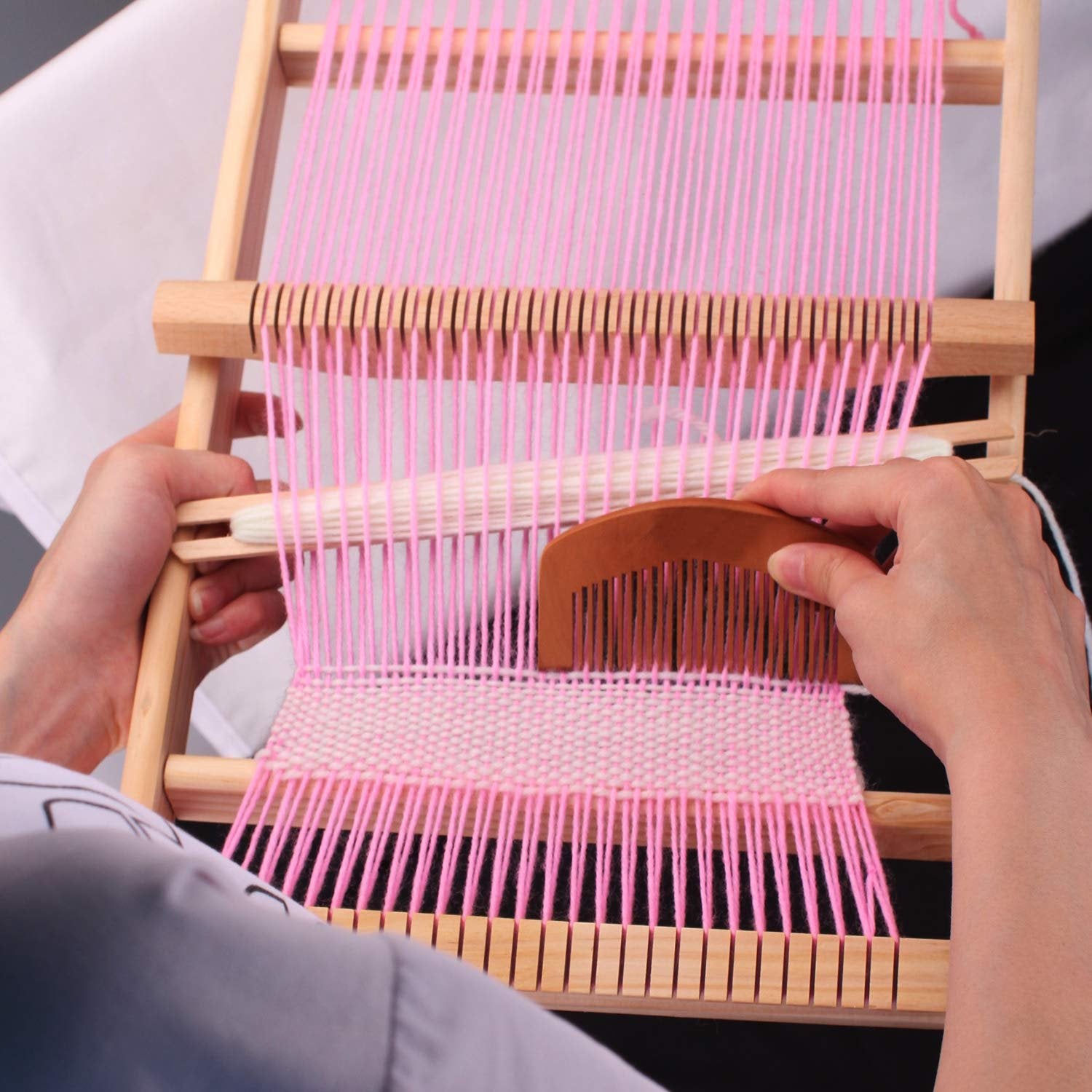 Hands weaving pink yarn on a wooden loom frame with a wooden comb tool.