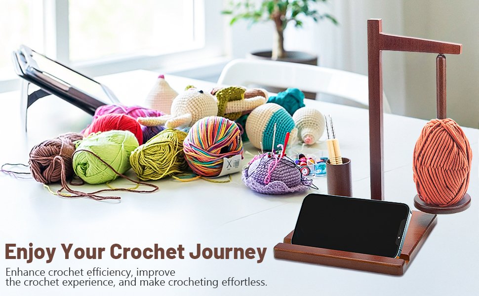 Wooden yarn holder with a ball of orange yarn, a phone stand, and various crochet supplies on a table.