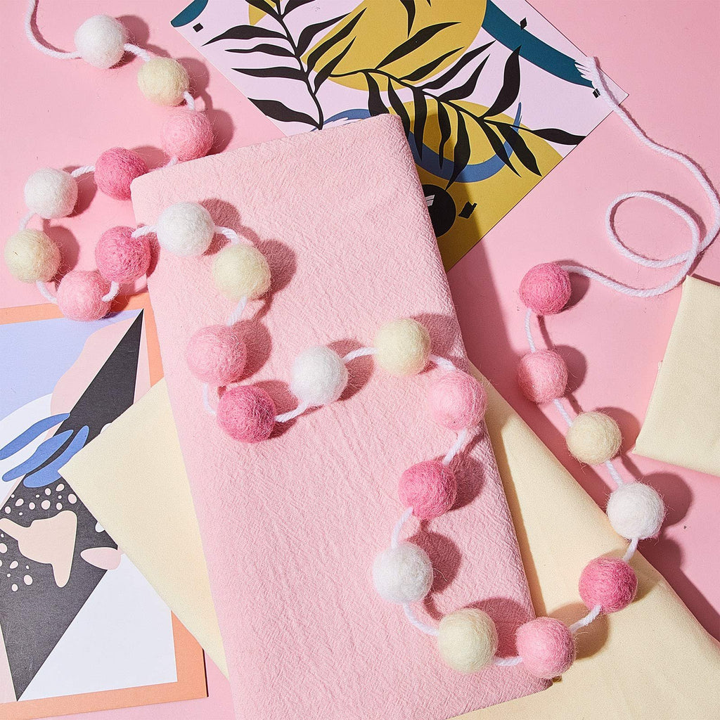 Two strands of pink, light pink, beige, and white wool felt pom pom garland draped over pink fabric and decorative cards.