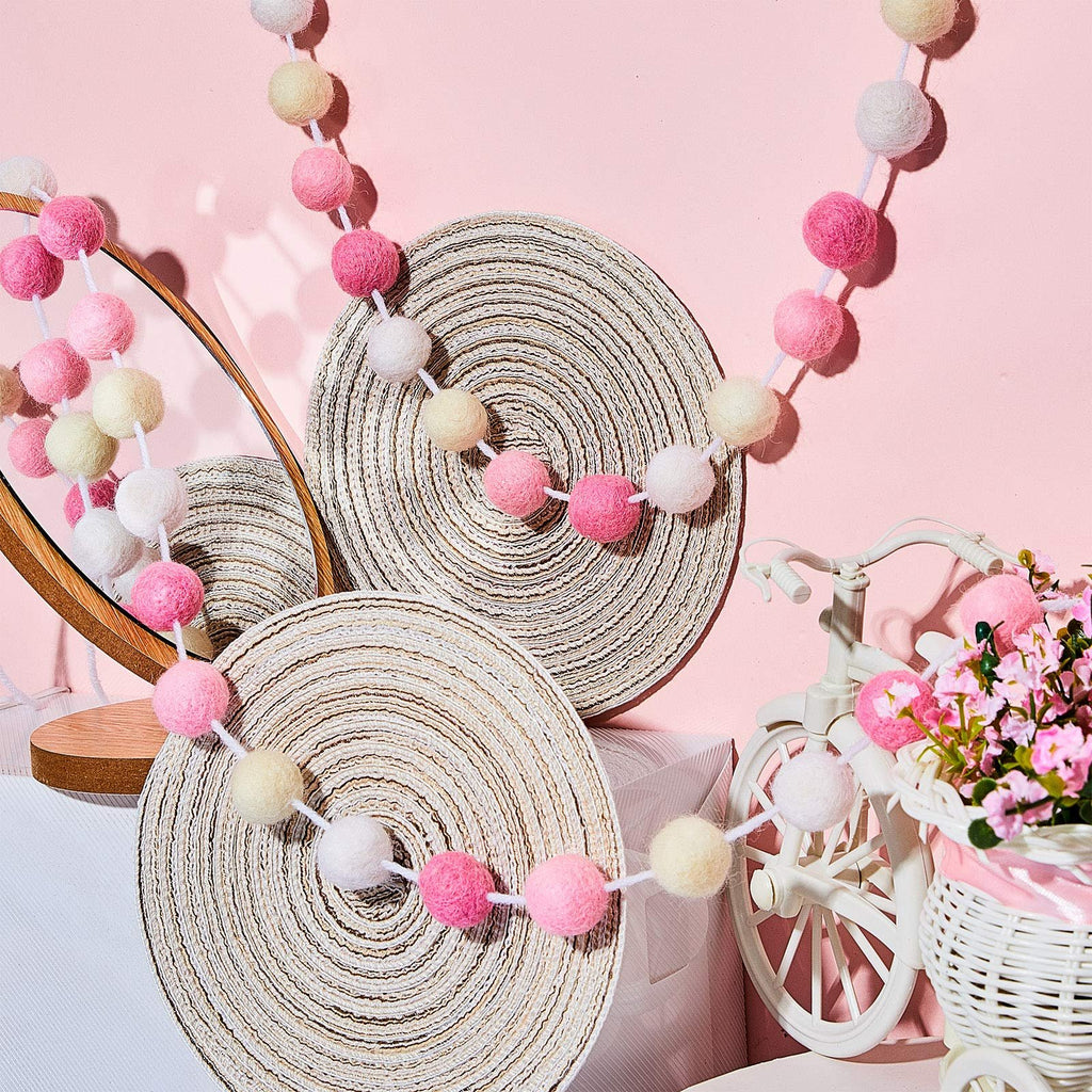 Wool felt pom pom garland in pink, light pink, beige, and white draped over decorative circular mats, with a mirror and a miniature bicycle with flowers.