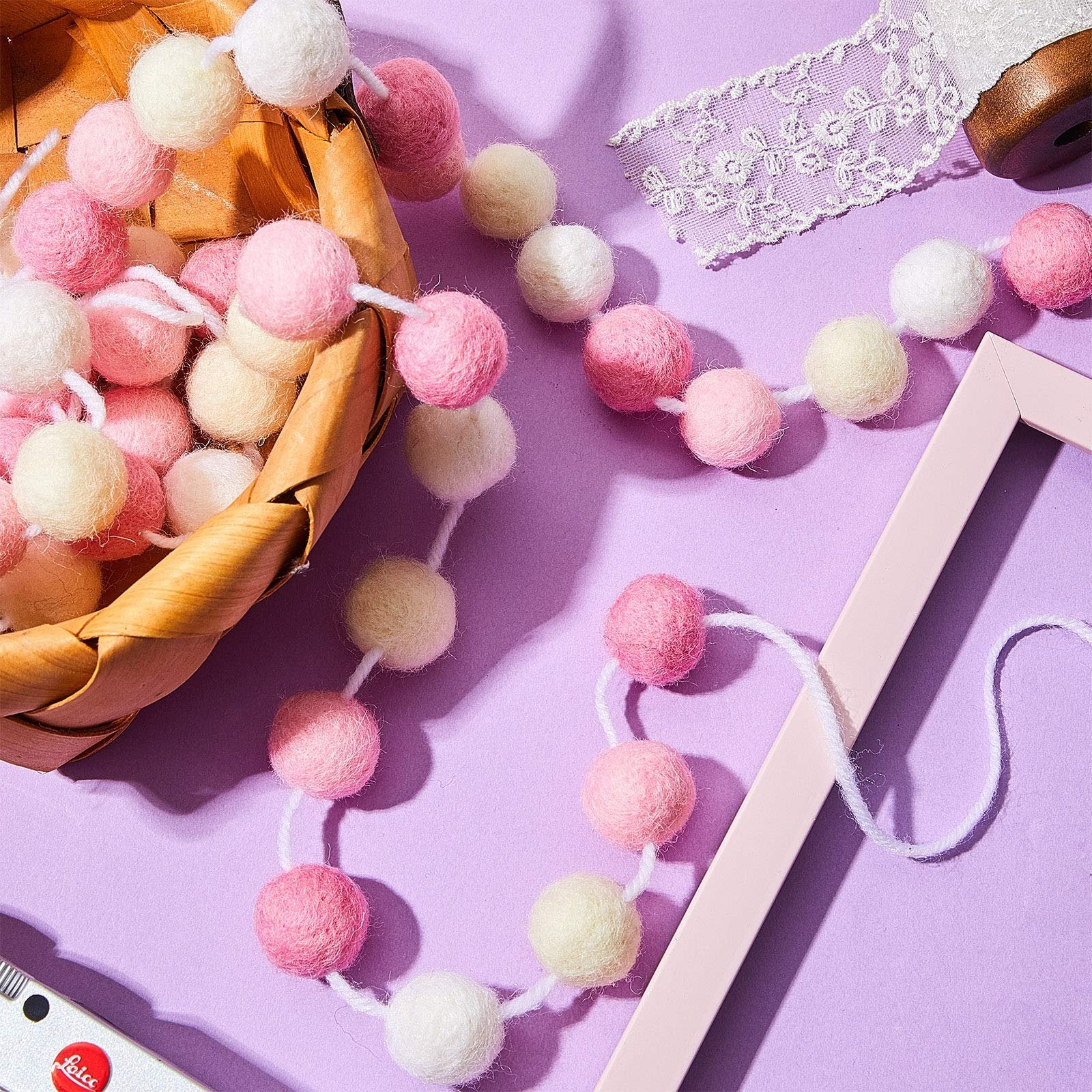 Wool felt pom pom garland in pink, light pink, beige, and white, spilling from a basket with lace and a pink frame.