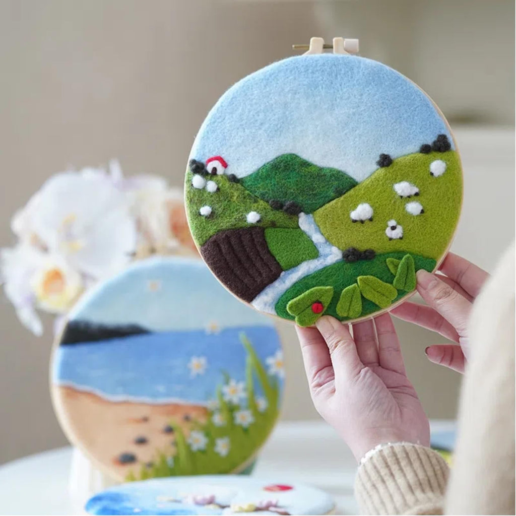 Hands holding a round needle-felted landscape art with sheep and a small house, with another felt art in the background.