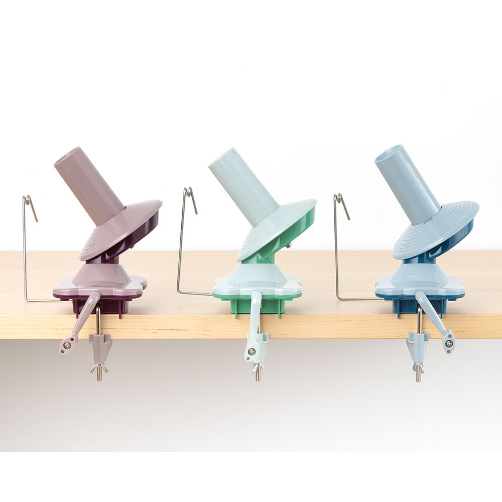 Three yarn ball winders in purple, green, and blue shades, clamped to a wooden shelf against a white background.