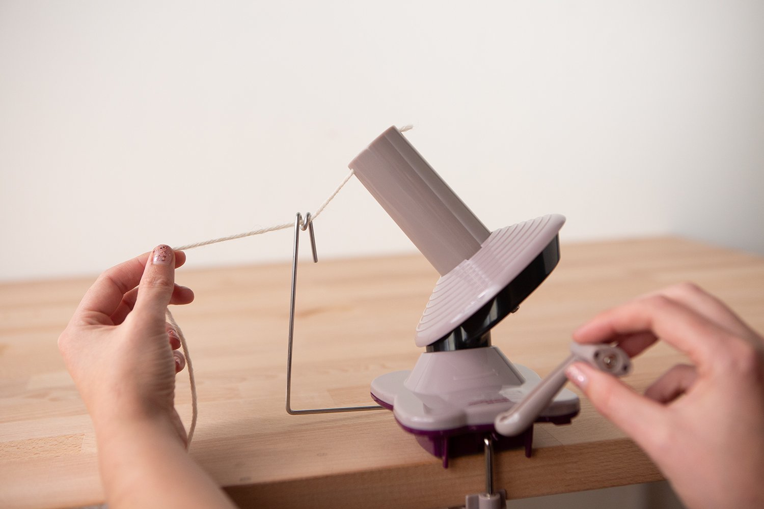 Hands using a purple yarn ball winder on a wooden table to wind white yarn.