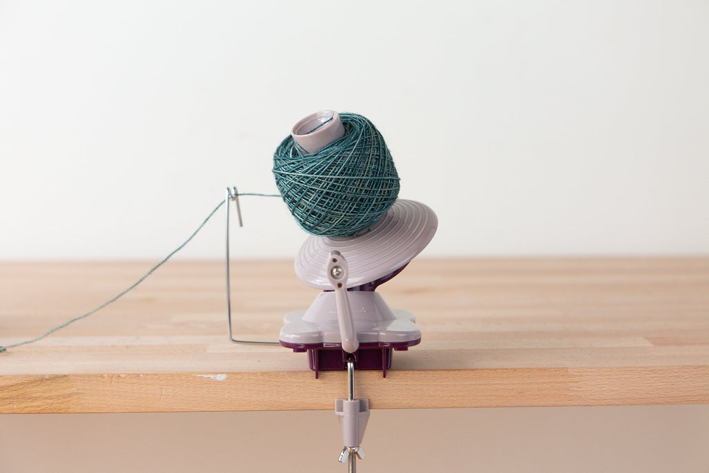 Purple yarn ball winder with teal yarn attached, clamped to a wooden surface.